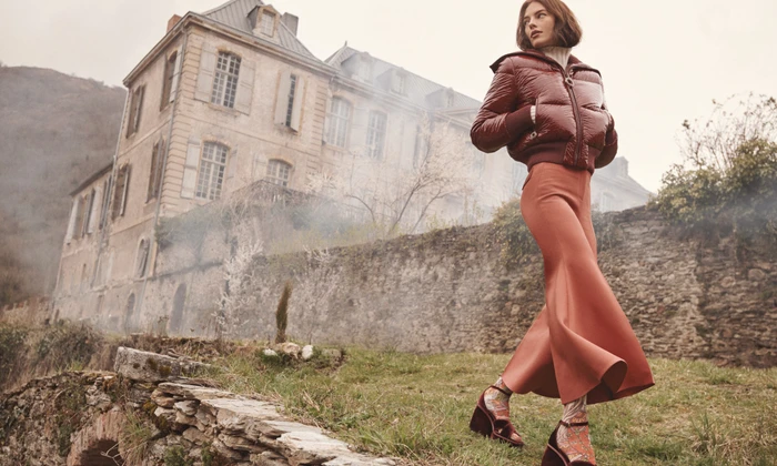 Model wearing a glossy dark red puffer jacket and terracotta skirt walking outdoors before a large stone building.