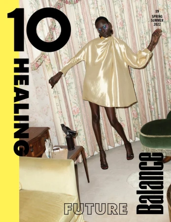 Model Ajak Deng wearing a metallic gold dress against patterned floral curtains and vintage furnishings.