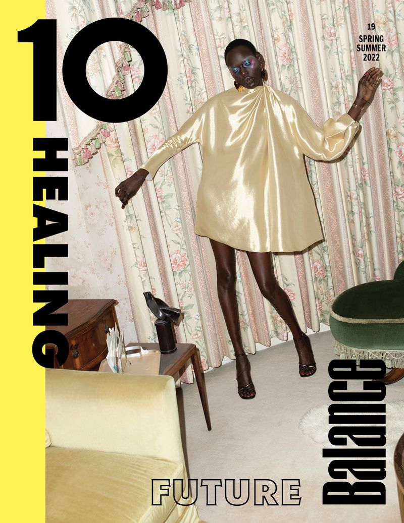 A dark-skinned fashion model in a shiny gold dress poses inside a room with vintage wallpaper, next to a yellow chair and side table.