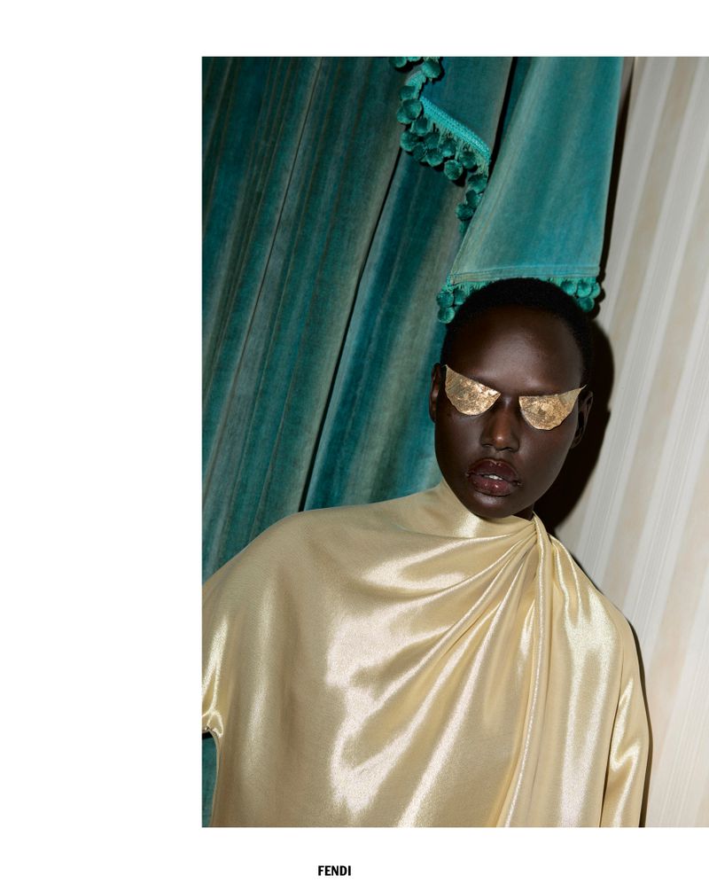 Fashion shot of a dark-skinned woman with gold foil sunglasses, wearing a shiny gold drape top, posing in front of a curtain.