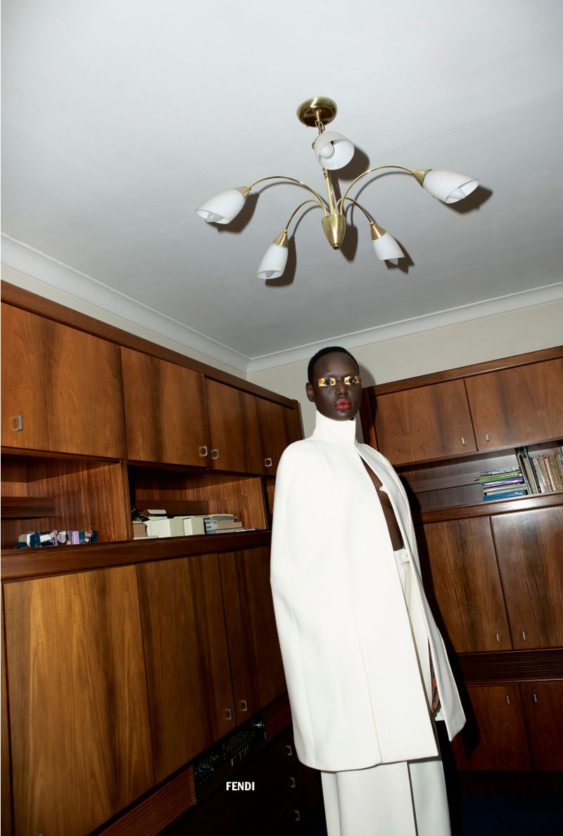 Elegant fashion shot indoors. A black female model wears an off-white cape and pants, standing against a cabinet, chandelier overhead.