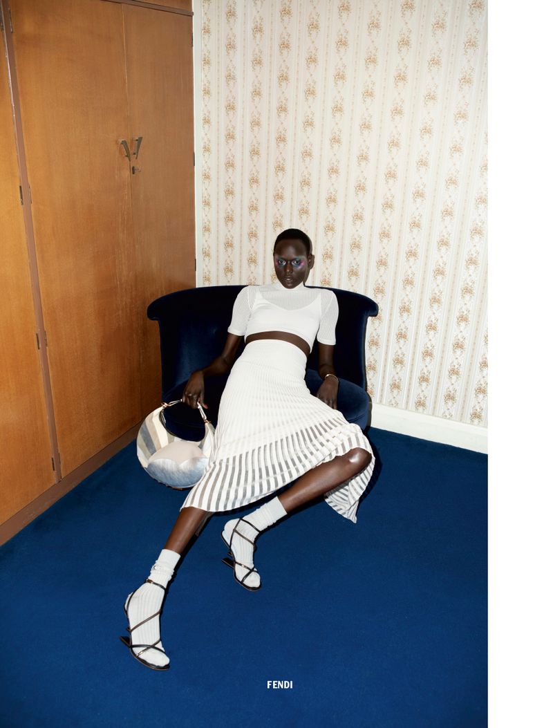 Fashion editorial shot of a Black woman in white knitwear sitting on a blue velvet chair, holding a silver handbag in an indoor setting.