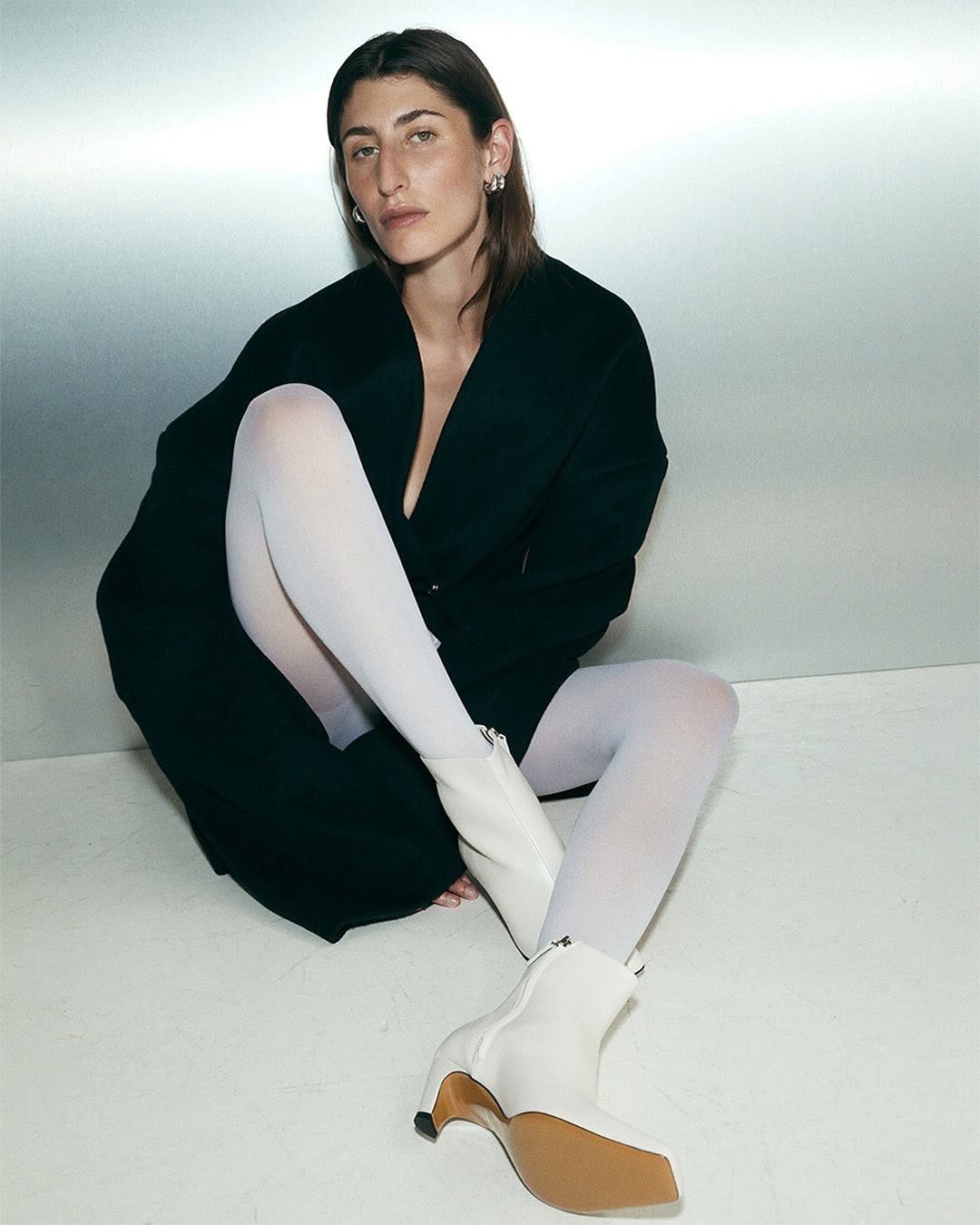 Model seated on the floor wearing a black coat, white tights, and white heeled boots against a metallic background