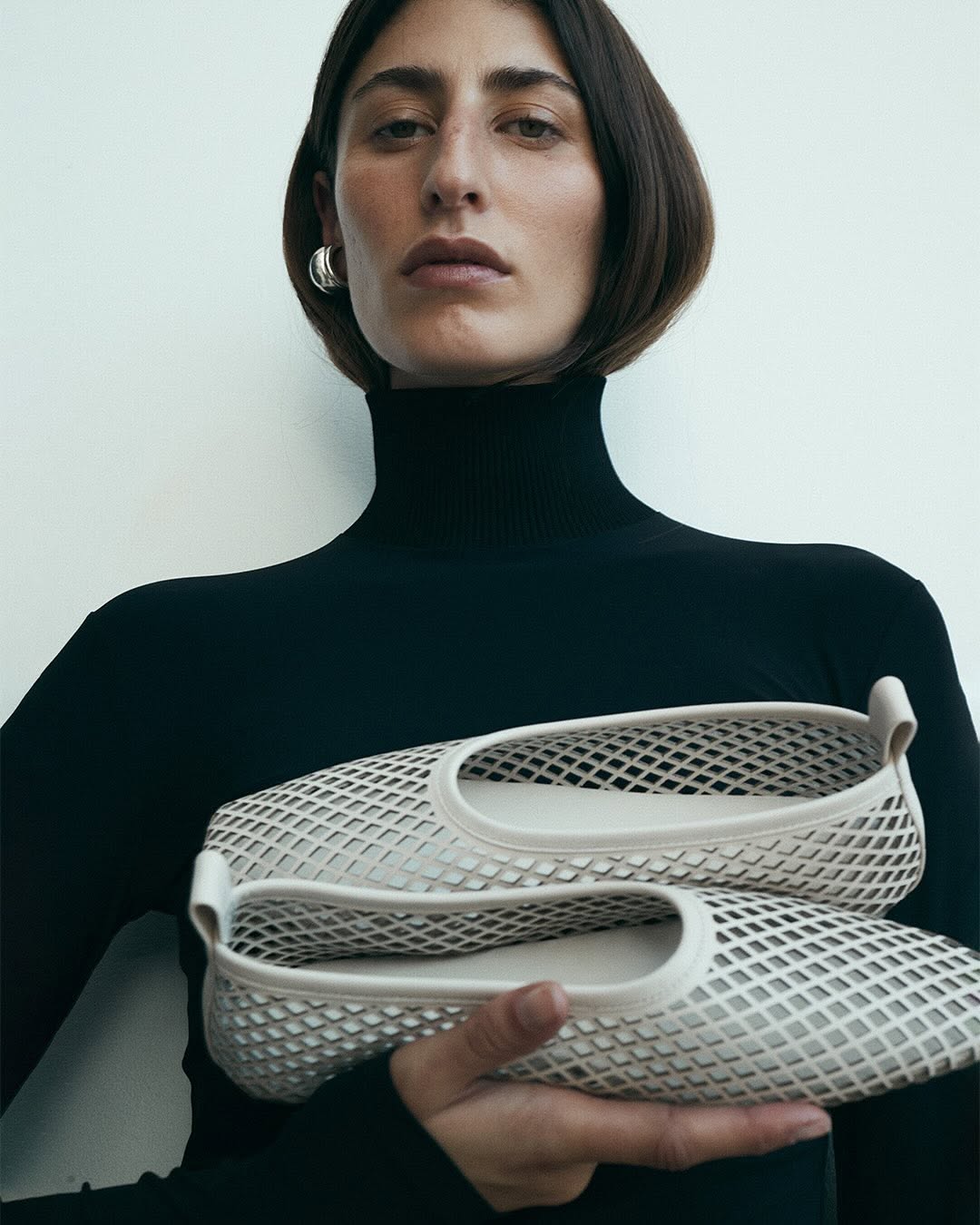 Woman in a black turtleneck holds a pair of cream-colored, lattice-patterned ballet flats in a studio setting with soft, neutral lighting.