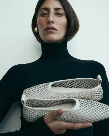 Woman in a black turtleneck holds a pair of cream-colored, lattice-patterned ballet flats in a studio setting with soft, neutral lighting.