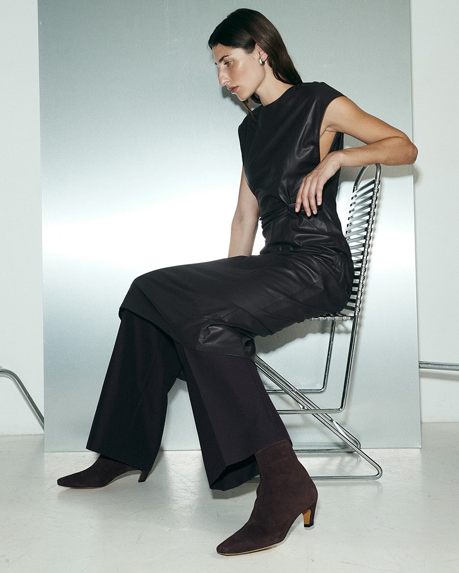 Elegant woman in black leather dress and brown ankle boots sits on a metal chair against a shiny silver background in a fashion setting.