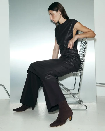 Elegant woman in black leather dress and brown ankle boots sits on a metal chair against a shiny silver background in a fashion setting.