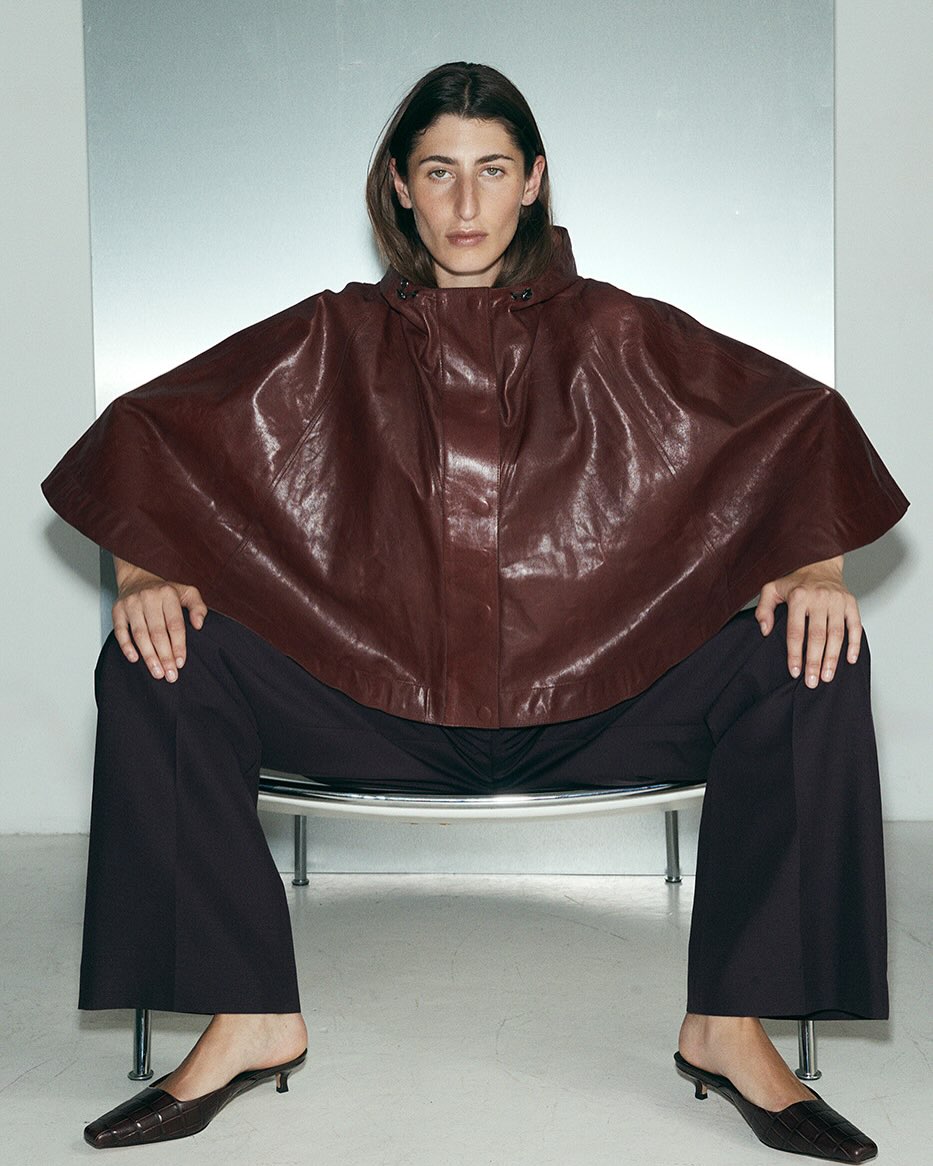 Elegant adult woman in a hooded burgundy cape and dark trousers, sitting on a wire chair against a grey background in a fashion shot.
