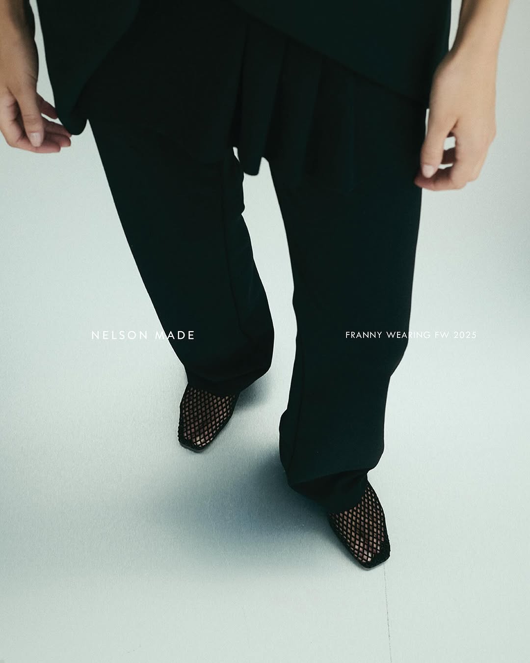 Overhead shot features model in black top and pants, showcasing black mesh heels on a light silver backdrop, emphasizing fashion details.