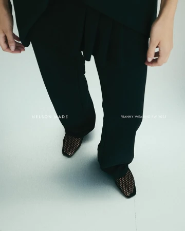Overhead shot features model in black top and pants, showcasing black mesh heels on a light silver backdrop, emphasizing fashion details.
