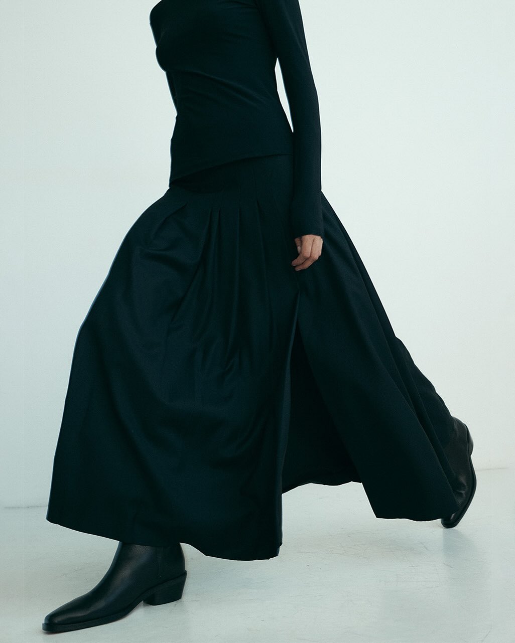 Fashion shoot of woman in long-sleeve shirt, long skirt with a slit, and ankle boots, walking in a bright, neutral-tone studio setting.