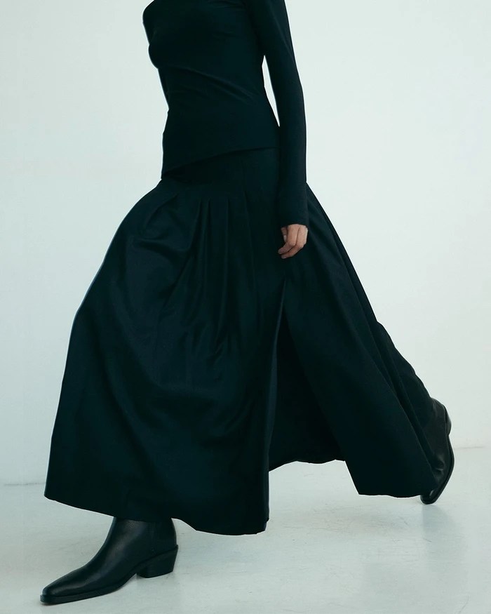 Model in dark top, long pleated skirt, and black boots walking against a plain white studio background.