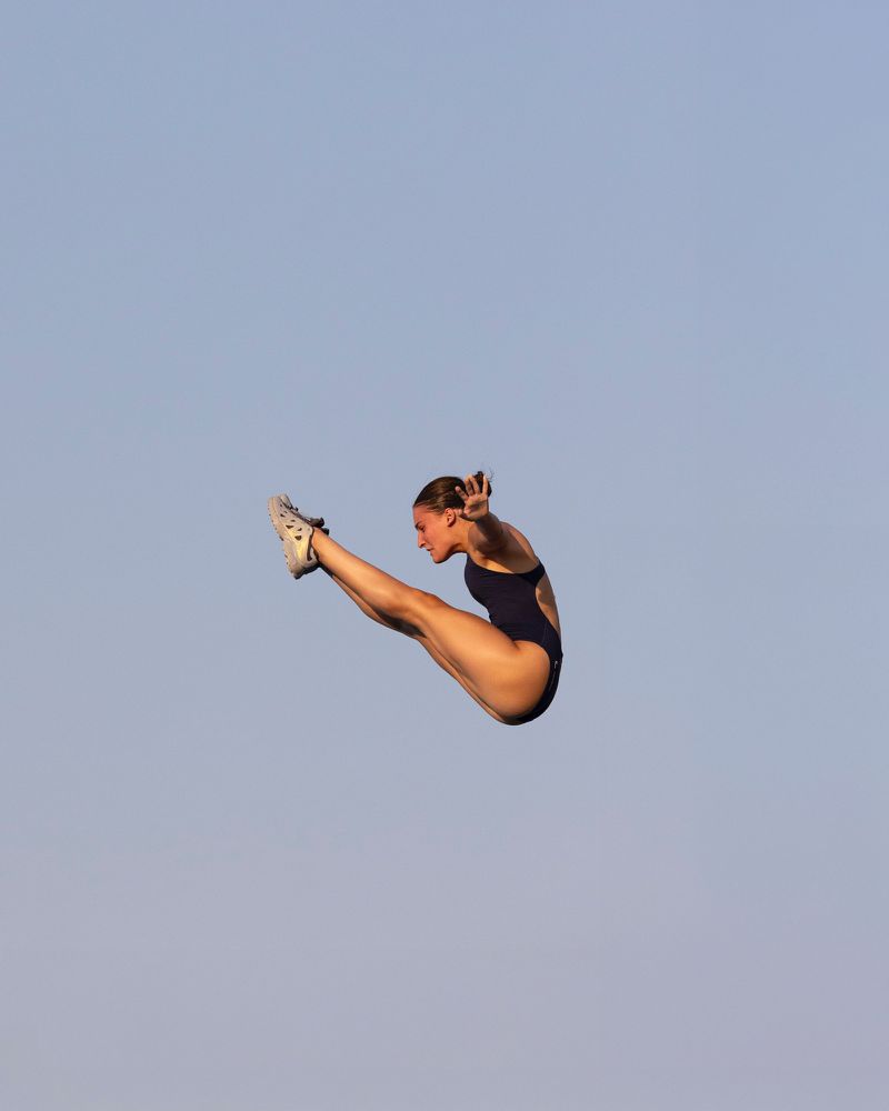 Female diver in mid-air with arms outstretched and legs together, poised against a clear blue sky, highlighting precision and athleticism.