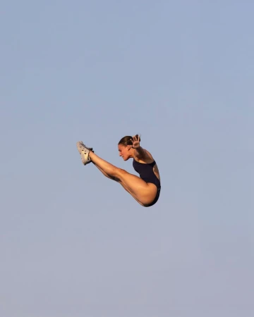 Female diver in mid-air with arms outstretched and legs together, poised against a clear blue sky, highlighting precision and athleticism.