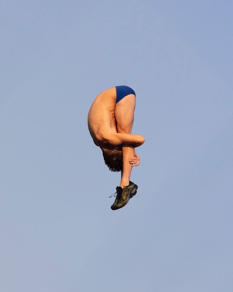 Man in mid-air performing a stunt, diving or acrobatic movement, wearing blue swimwear and shoes against a clear blue sky backdrop.