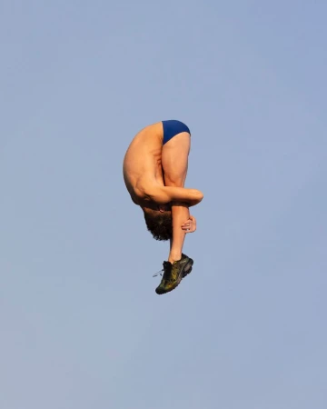 Man in mid-air performing a stunt, diving or acrobatic movement, wearing blue swimwear and shoes against a clear blue sky backdrop.