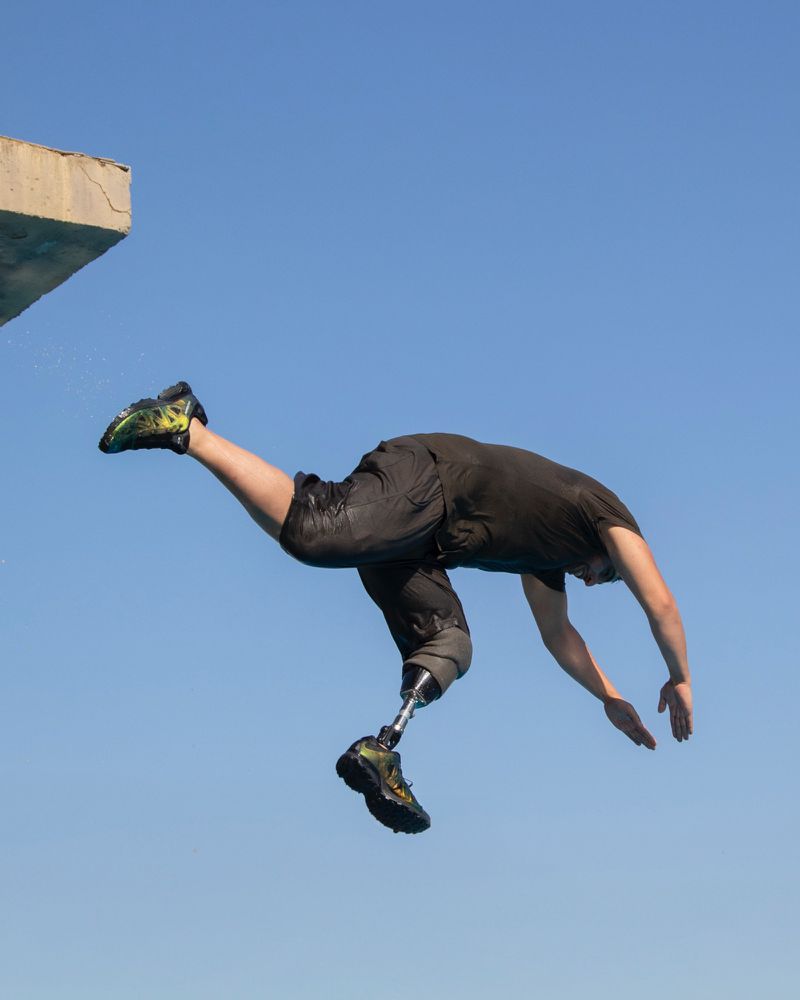Adult male dives into a pool. He has a prosthetic leg. He is wearing a black top and shorts. The sky and water are blue.