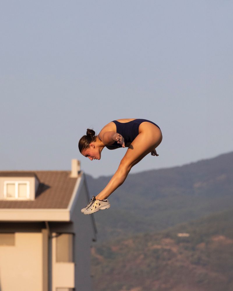 A young female athlete wearing a navy swimsuit performs a dive, captured at eye-level in an outdoor setting with a clear sky.