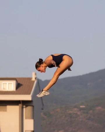 A young female athlete wearing a navy swimsuit performs a dive, captured at eye-level in an outdoor setting with a clear sky.