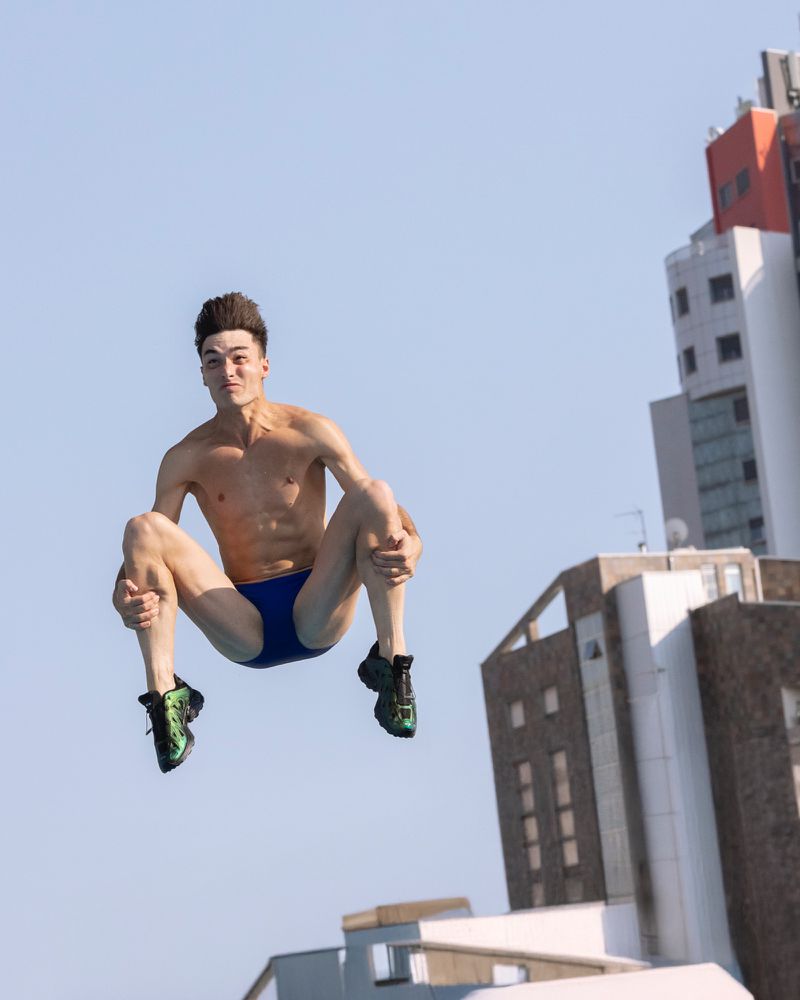 Male diver leaps from an unseen platform, body tucked, against a backdrop of bright sky and modern buildings in an urban environment.