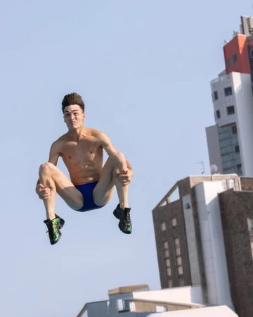 Male diver leaps from an unseen platform, body tucked, against a backdrop of bright sky and modern buildings in an urban environment.