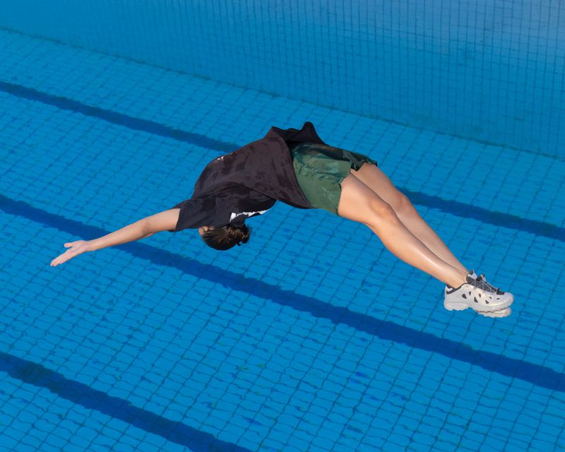 An athlete arches over an outdoor swimming pool, captured from above. The swimmer is wearing a black shirt and green shorts.