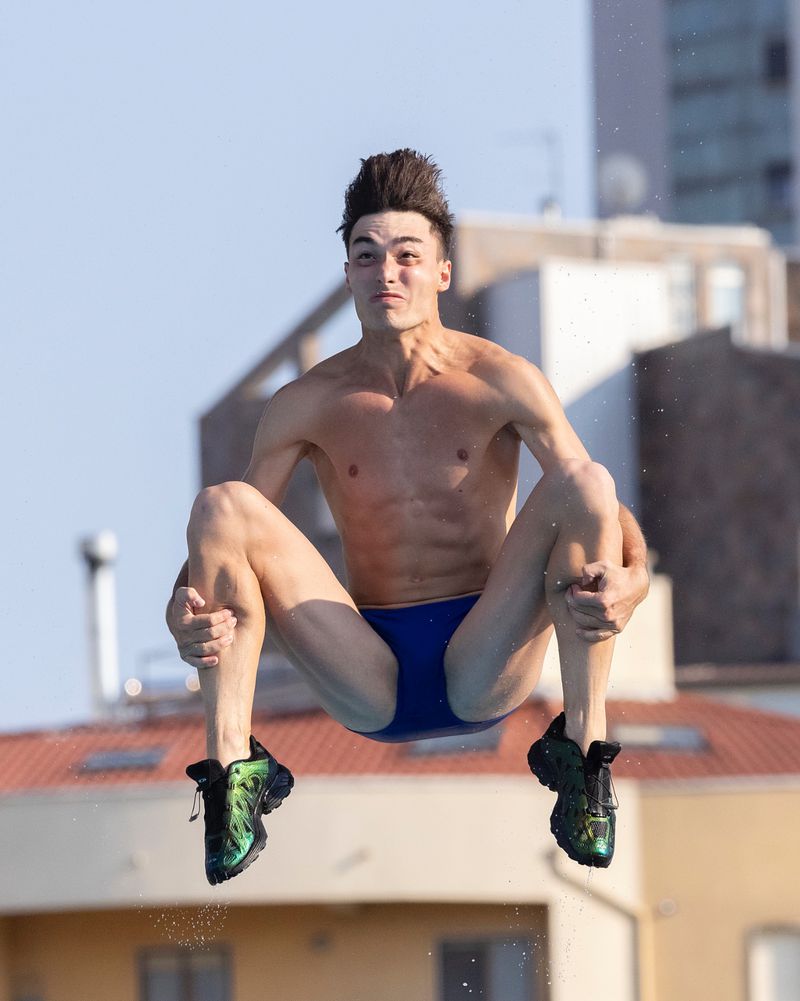 Male diver captured mid-air in a dynamic pose against a blue sky and urban backdrop, wearing a blue swimsuit and colorful shoes.