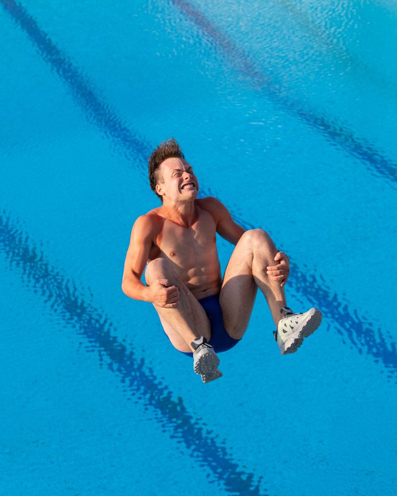 Athlete in mid-air above a pool, showcasing a dramatic dive. The diver is captured against a vivid blue pool backdrop with dynamic composition.