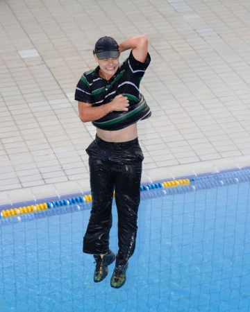 Young man with cap, black pants and t-shirt removes his t-shirt while jumping into swimming pool with blue water and white tile border.