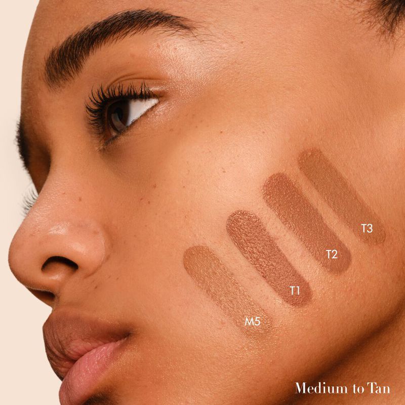 Close-up of a tan-skinned adult woman's face with four brown makeup swatches on her cheek, labeled M5, T1, T2, and T3.