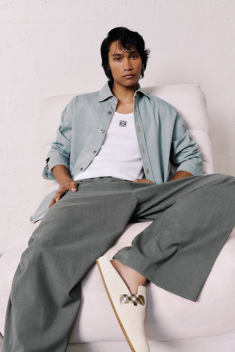 Man in casual outfit with shirt, white top, and wide-leg pants, seated indoors. Beige loafers complete the relaxed, fashionable look.