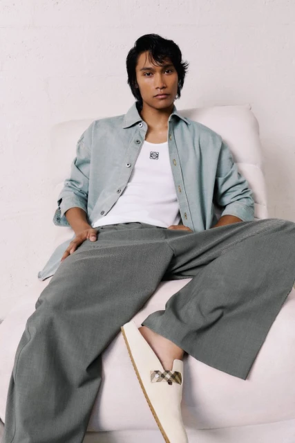 Man in casual outfit with shirt, white top, and wide-leg pants, seated indoors. Beige loafers complete the relaxed, fashionable look.