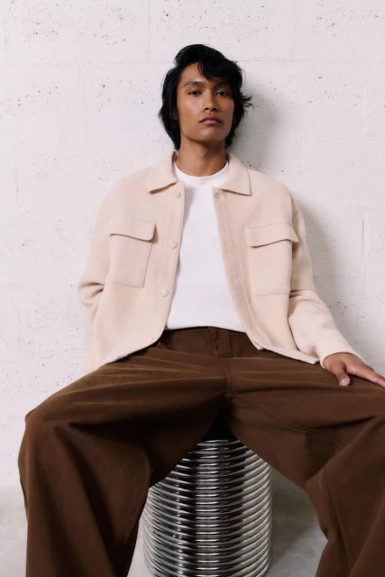 A light-skinned male model poses sitting indoors on a metal stool, wearing a beige shirt jacket over a white t-shirt and brown slacks.