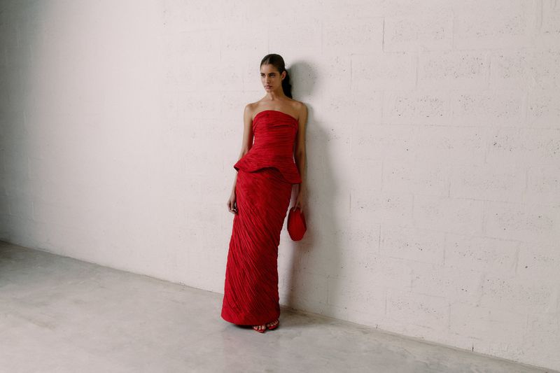 A woman in a red, floor-length gown stands against a white wall, holding a matching clutch. Minimalist fashion photography.
