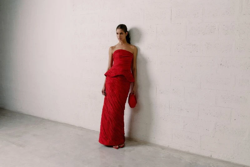 A woman in a red, floor-length gown stands against a white wall, holding a matching clutch. Minimalist fashion photography.