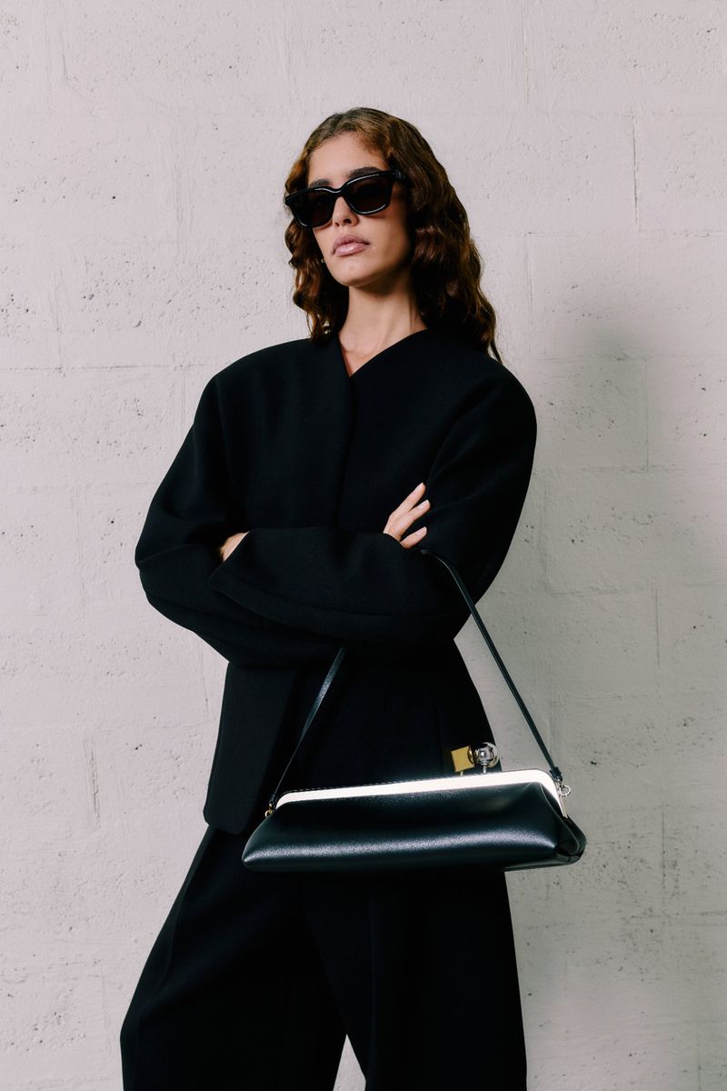 Elegant woman in black jacket and wide-leg pants, posing with a black frame bag against a textured wall, sunglasses on, fashion forward.