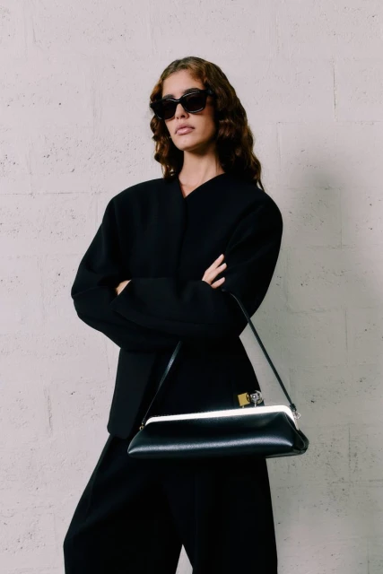 Elegant woman in black jacket and wide-leg pants, posing with a black frame bag against a textured wall, sunglasses on, fashion forward.