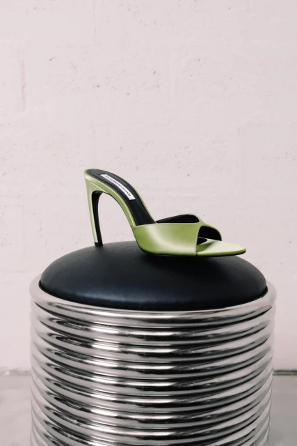 Elegant lime-green heeled sandal with a black insole sits on a round, textured silver and black stool against a pale off-white wall.