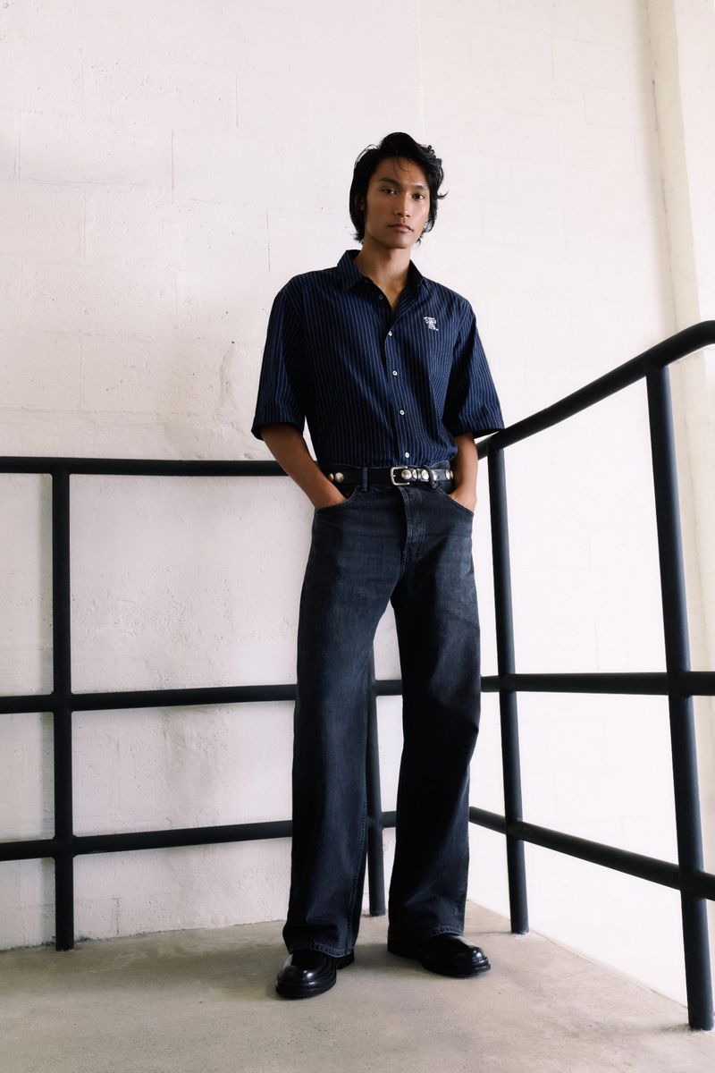 Model wearing a navy striped short-sleeve shirt and dark wide-leg jeans standing by black metal railings against a light wall.