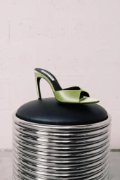 Light green satin high-heeled mule displayed on a round black cushioned stool with a ribbed chrome base.
