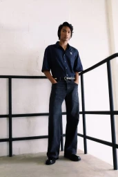 Model wearing a navy striped short-sleeve shirt and dark wide-leg jeans standing by black metal railings against a light wall.