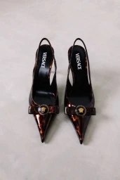 A pair of pointed-toe tortoiseshell slingback heels with bow details and circular gold emblems on a light grey background.