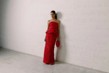 Model posing in a strapless red peplum maxi dress with a matching red clutch bag against a light textured wall.