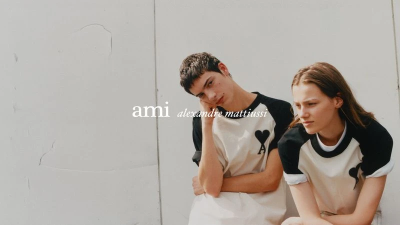 Two young adults, a male and a female, pose in matching white and black shirts in front of a textured white wall with diffused lighting.