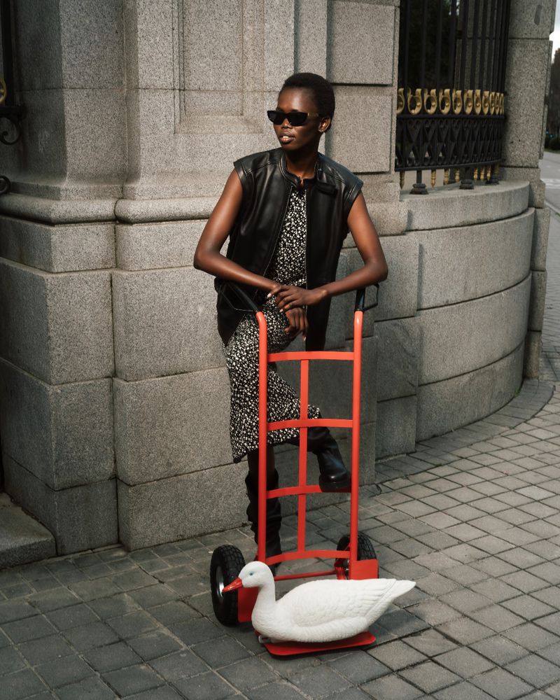 Stylish dark-skinned woman with sunglasses and black vest poses with a red hand truck and a white duck in an urban outdoor setting.