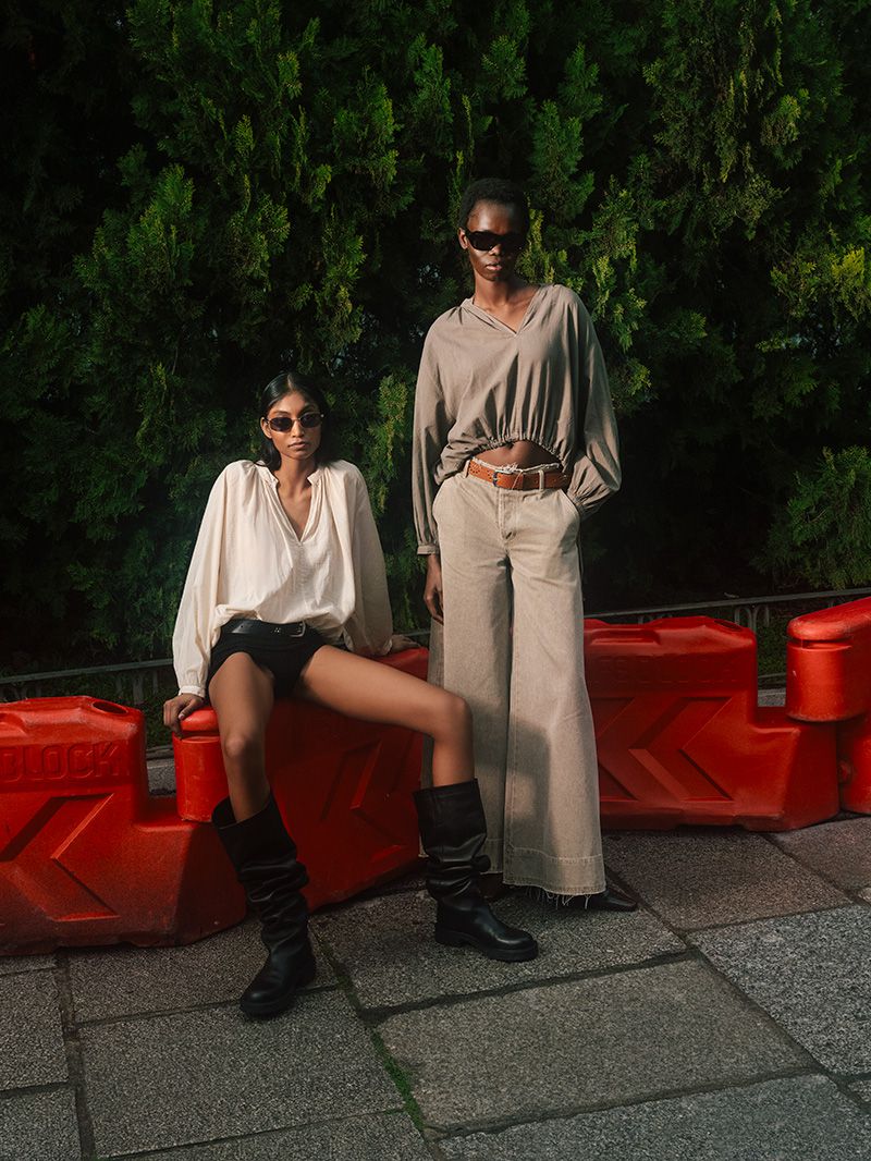 Two women pose in front of a green leafy hedge wearing trendy beige and white outfits with long sleeves and black leather boots.