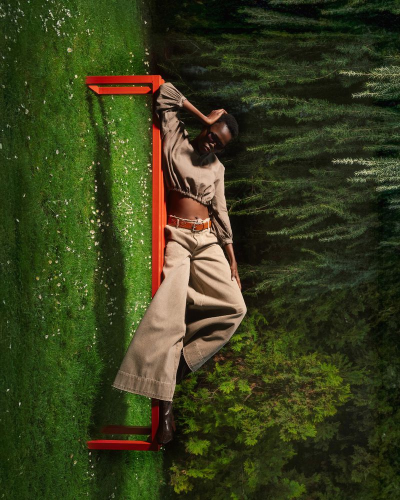 A woman in stylish tan outfit posing outside on a green lawn with trees, leaning against a red metal structure in an outdoor fashion shot.