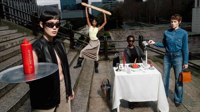 A fashion editorial photograph shows four women on outdoor steps with an unconventional outdoor dining scene featuring a baguette and ketchup.