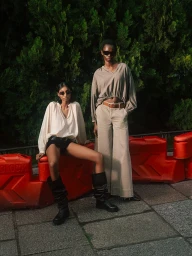 Two models wearing sunglasses, neutral tops, and wide-leg trousers posing against green foliage by a red barrier for MAX&Co.