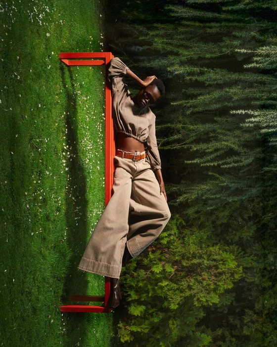 Model posing against a bright red bench outdoors in a garden setting wearing neutral-toned apparel.